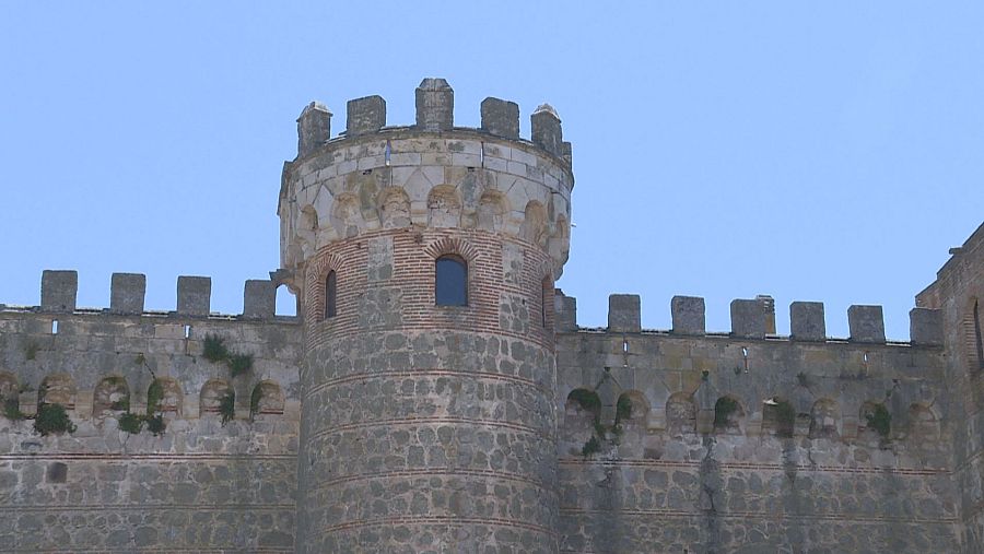 Torre circular de piedra gris con almenas, banda de ladrillos y pequeña ventana.  Muralla adjunta con arcos y almenas cuadradas; vegetación presente.