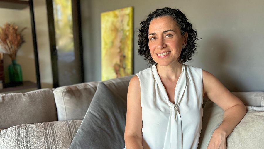 Retrato de Silvina Cornillón sonriendo, sentada en un sofá claro con blusa blanca sin mangas. Fondo con pared gris y detalles decorativos.