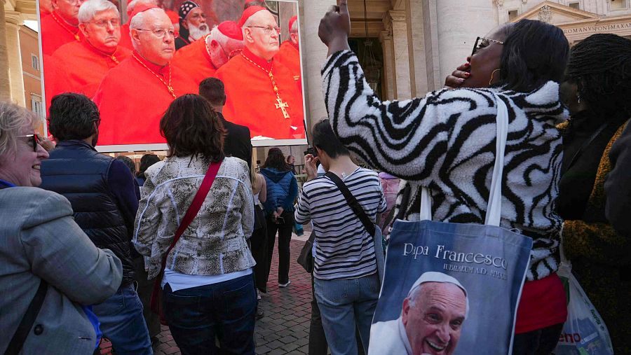 Una asistente con una bolsa que representa al difunto papa Francisco mientras observa una pantalla gigante que muestra imágenes de la santa misa de cardenales a la capilla Sixtina