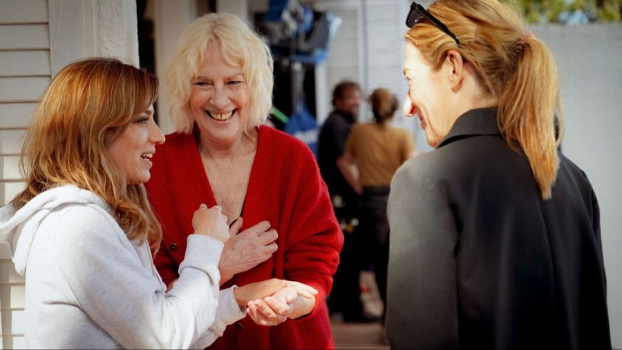 Tres mujeres sonríen en un set de rodaje; una lleva sudadera blanca, otra chaqueta roja y la tercera una chaqueta oscura.