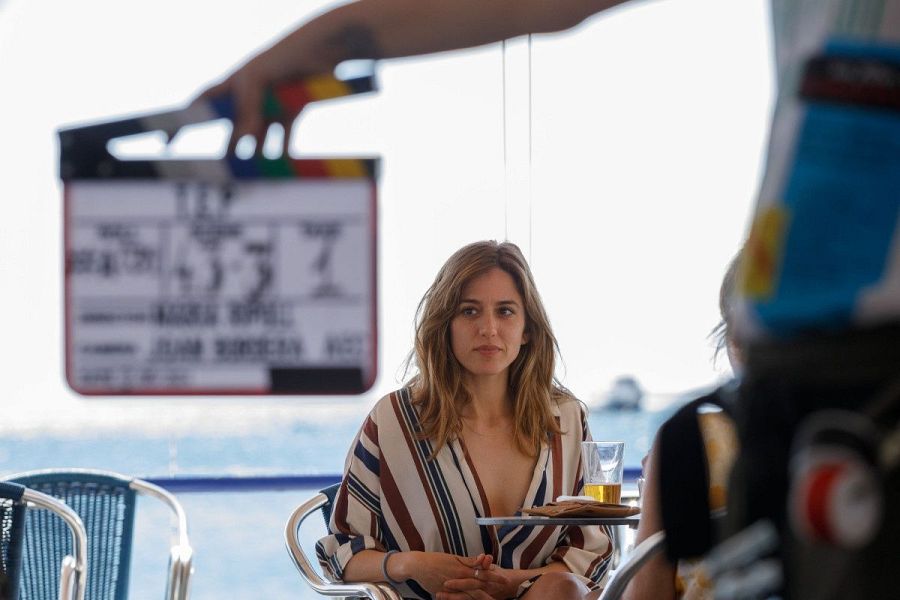 Actriz sentada en terraza con vistas al mar, expresión seria, camisa a rayas, claqueta de cine en primer plano, rodaje de película.