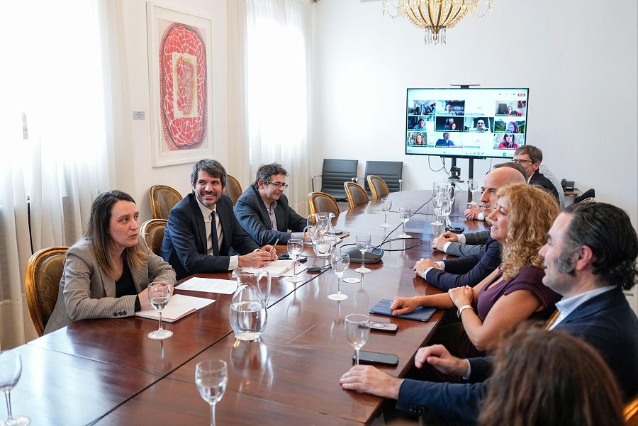 Reunión del ministro de Cultura con participantes presenciales y remotos.  Se observa una mesa con objetos varios y una pantalla mostrando una videoconferencia.