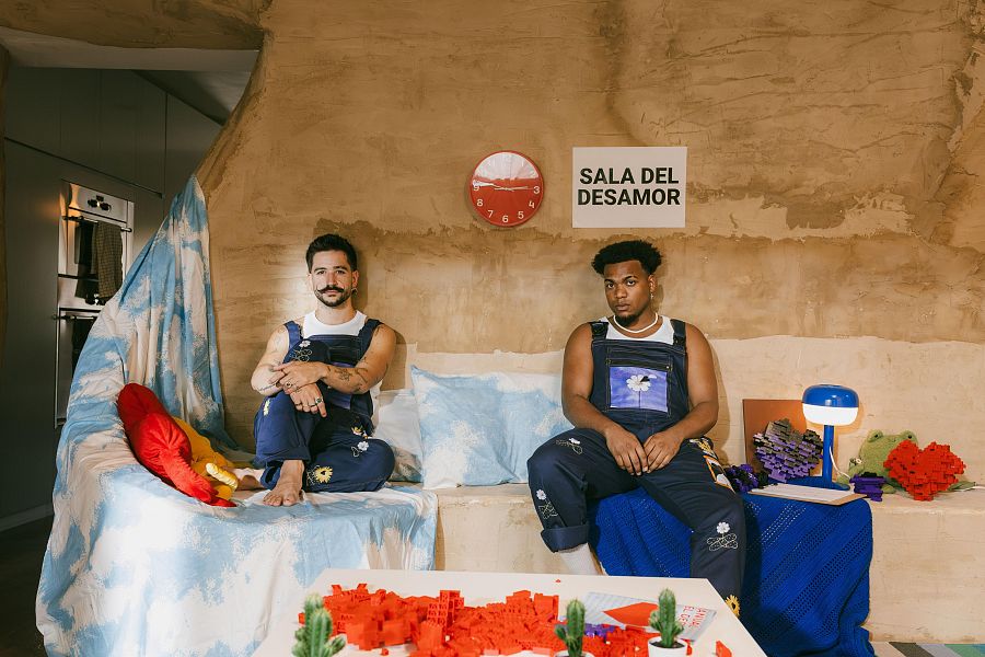 Dos artistas, sentados en un sofá con piezas de Lego, corazones y otras figuras a su alrededor, en un espacio decorado con un reloj rojo y un cartel. Ambiente reflexivo.