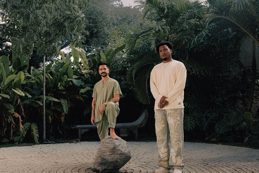 Dos músicos, uno en verde oliva y otro con jersey crema y vaqueros, posan en un jardín tropical.  Colaboración musical.