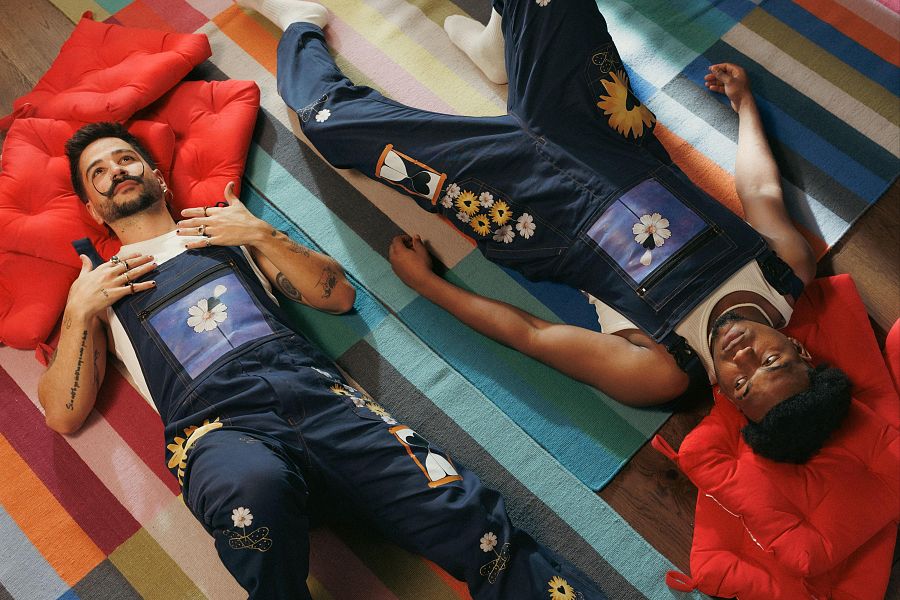 Dos hombres con overoles de mezclilla bordados descansan sobre una alfombra multicolor.  Ambiente tranquilo y reflexivo.