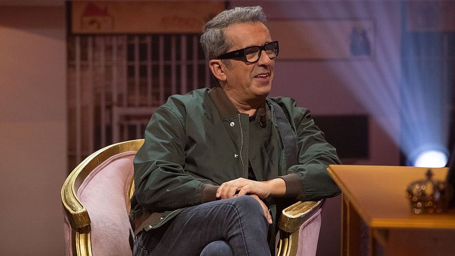 Andreu Buenafuente, con chaqueta verde y gafas, sentado en un sillón rosa, piernas cruzadas. Fondo con reja y corona dorada.