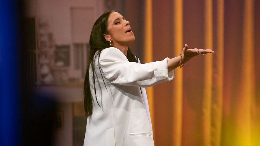 María Peláe canta con pasión en un escenario con cortinas doradas, luciendo una chaqueta blanca.  Imagen de una actuación en vivo.
