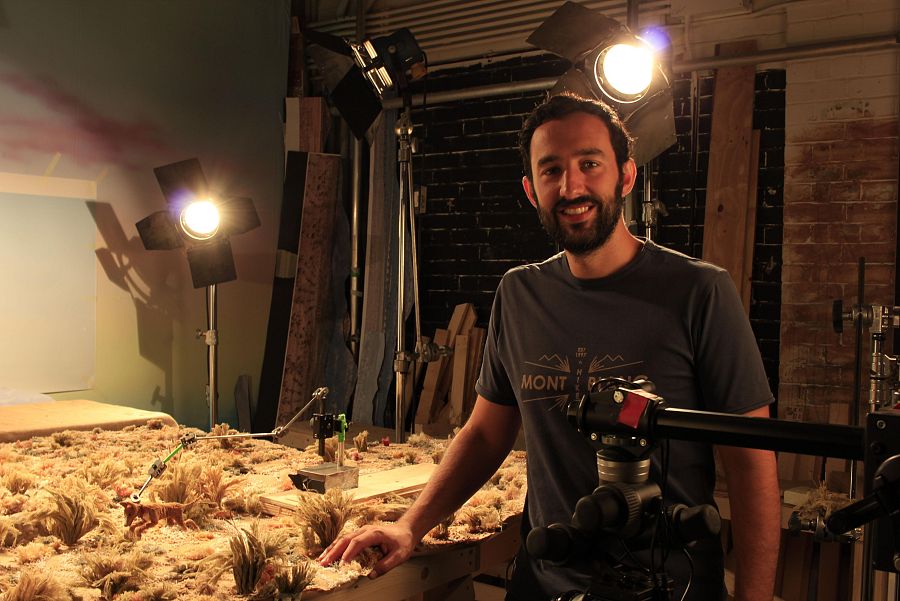 José Prats posa sonriente junto a una maqueta de paisaje desértico en un estudio de grabación, con equipos de filmación visibles.