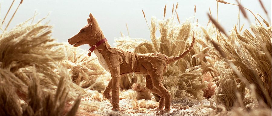 Perro animado de papel o tela marrón, con collar rojizo, en paisaje árido con plantas altas. Textura cosida o remendada.