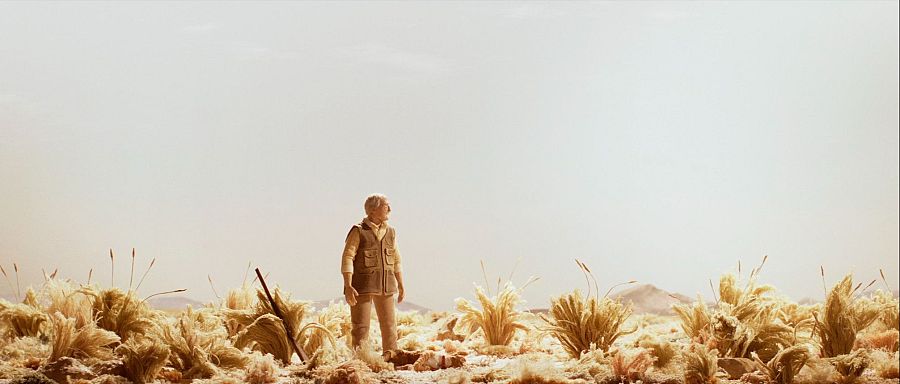 Hombre mayor en paisaje desértico, chaleco marrón, pantalones beige, escopeta, actitud contemplativa. Fotograma de animación.