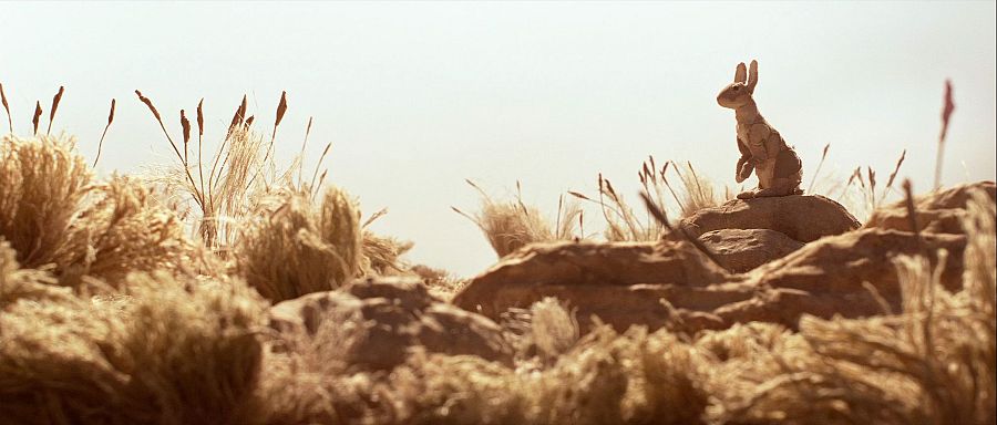 Conejo de animación marrón claro sentado en roca desértica, mirando a la derecha.  Paisaje árido, cielo soleado.