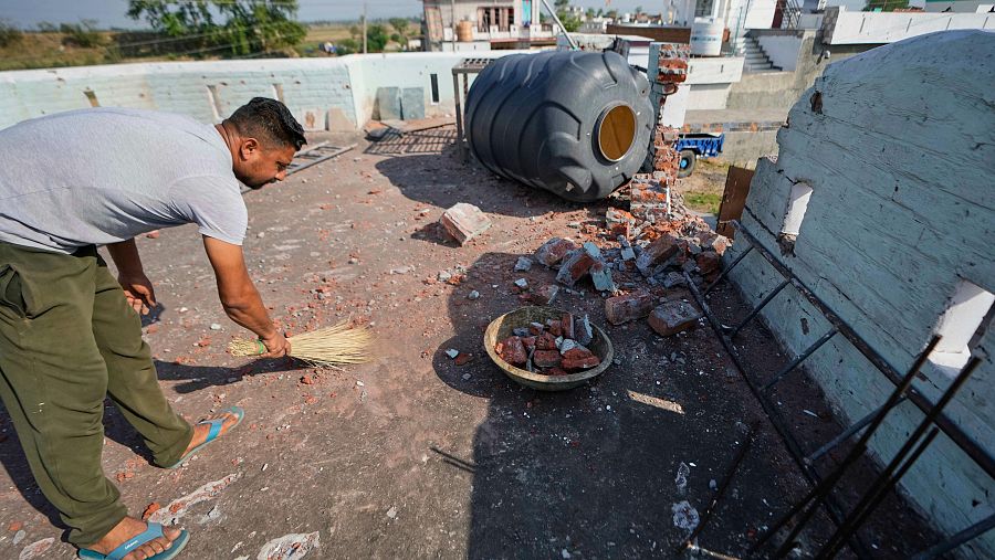 Hombre limpiando escombros de azotea tras ataques aéreos.  Ladrillos rotos, tanque de agua volcado y pared dañada muestran la destrucción.