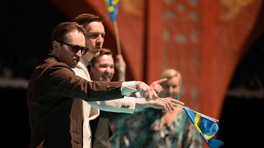 Tres hombres en trajes, con banderas suecas, en un evento; uno señala, otro parece cantar. Fondo desenfocado con una mujer y otra bandera.
