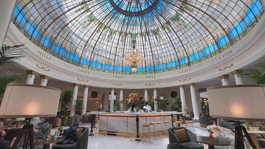 Impresionante cúpula de cristal con secciones azules y detalles vegetales, salón con barra de mármol blanco, mesas y asientos en tonos grises y verdes, iluminación cálida y detalles arquitectónicos en blanco y dorado.
