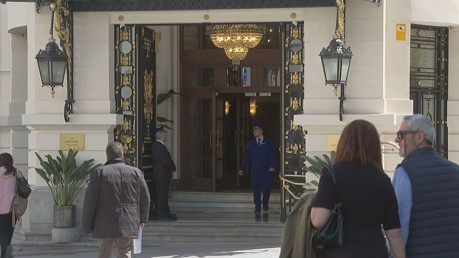 Entrada majestuosa del Hotel Palace con puertas de madera, portero uniformado y personas en la calle.  Gran lámpara de araña visible en el vestíbulo.