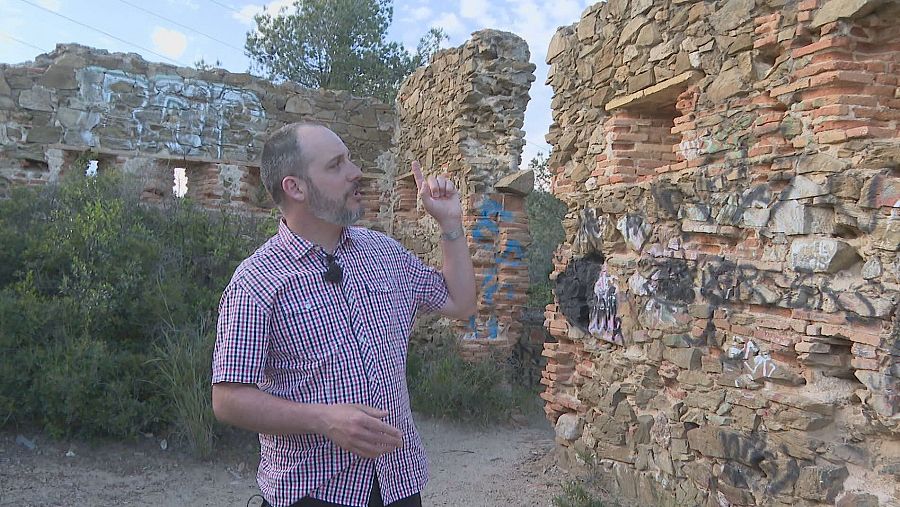 Historiador frente a una pared de piedra parcialmente destruida, con grafitis, explicando detalles con un micrófono inalámbrico.