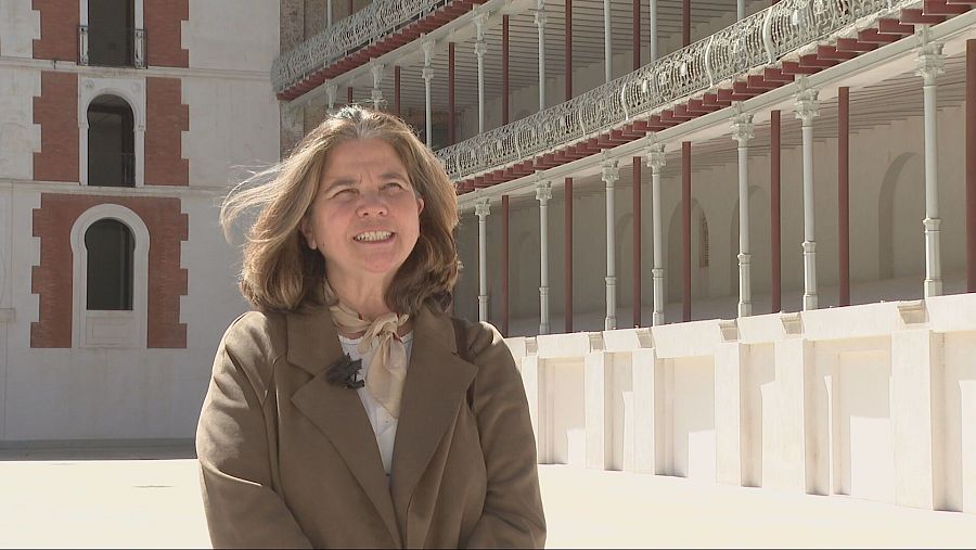 Arquitecta con abrigo marrón y bufanda beige, micrófono de solapa, en cancha de frontón con gradas y estructura visible al fondo.