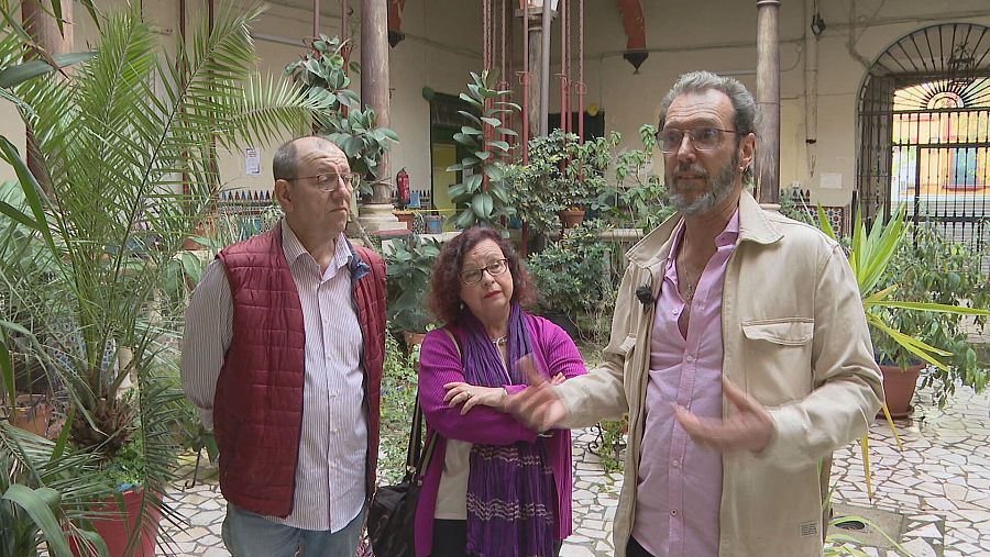 Patio interior con columnas y suelo embaldosado, tres personas conversan; una lleva chaleco granate, otra un jersey y pañuelo morado, y la tercera una chaqueta beige.