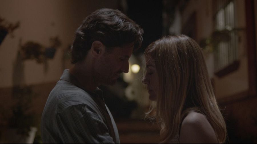 Pareja a punto de besarse en habitación con poca luz; él viste camisa oscura, ella tiene cabello rubio.  Decoración con figuras animales y ventana con barrotes al fondo.