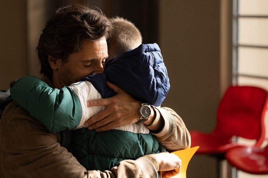 Abrazo emotivo entre un hombre con chaqueta marrón y un niño con chaqueta verde y blanca. El hombre lleva un reloj y sostiene un papel amarillo.  Escena conmovedora en un espacio público.