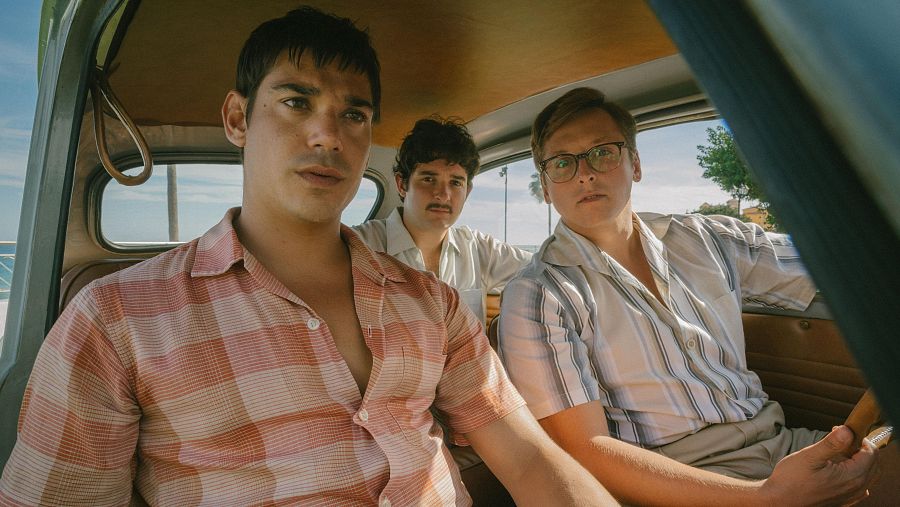 Tres jóvenes en un coche antiguo, uno conduce y los otros dos miran hacia adelante con expresiones serias.  Paisaje costero visible por las ventanas.