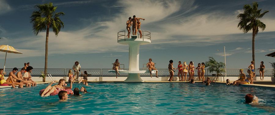 Escena de piscina soleada con cuatro personas en un trampolín, listas para saltar.  Mucha gente disfruta del agua y el ambiente veraniego.