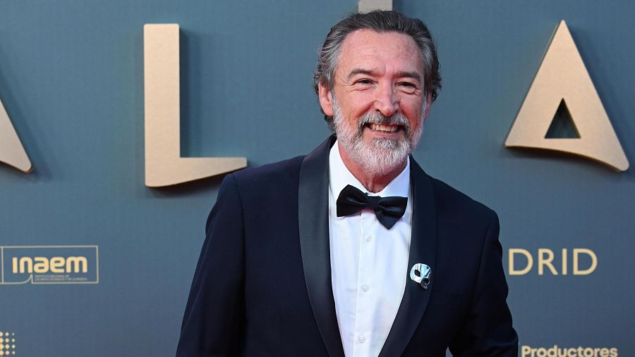 Actor con esmoquin negro y pajarita, sobre fondo azul con letras doradas 