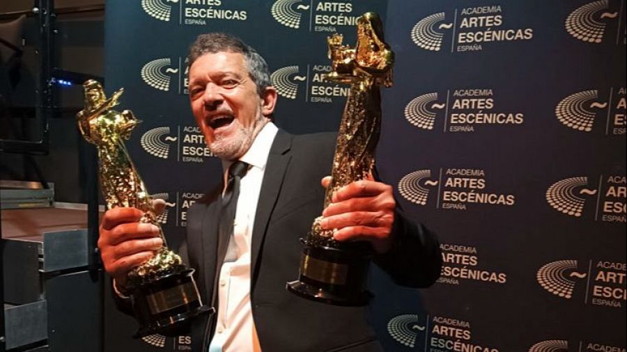 Antonio Banderas, sonriente, muestra dos premios dorados tras recibir un reconocimiento a su carrera. Lleva traje oscuro, camisa blanca y corbata negra.