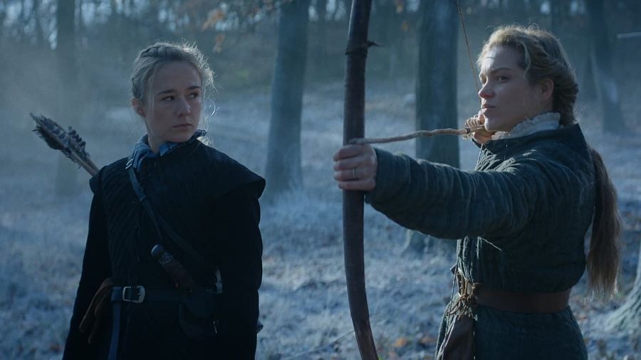 En un bosque nevado, dos mujeres con atuendos oscuros, una con ballesta y otra con arco y flecha tensado, protagonizan una escena de atmósfera fría y misteriosa.