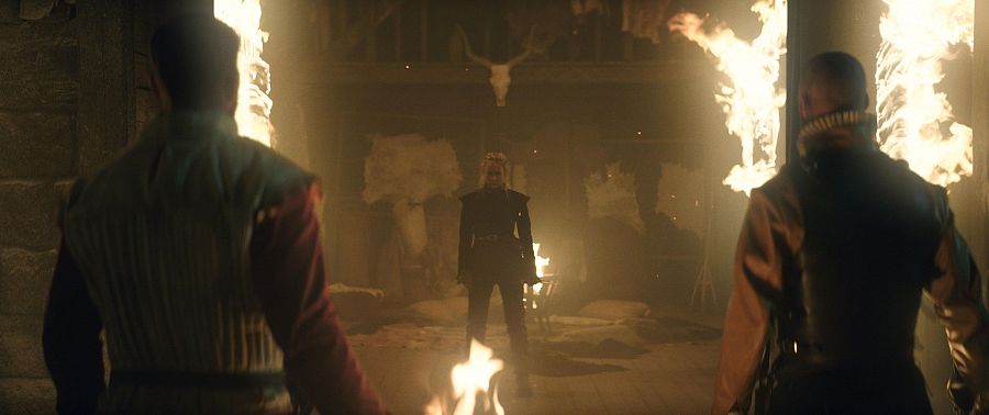 Escena dramática en un pasillo de piedra iluminado con antorchas. Una figura central en negro, flanqueada por otras dos, crea una atmósfera de tensión.