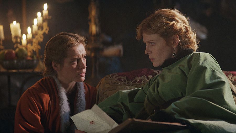 Escena interior nocturna: dos mujeres con vestidos de época, una con una carta, muestran expresiones serias y preocupadas bajo la luz de velas.