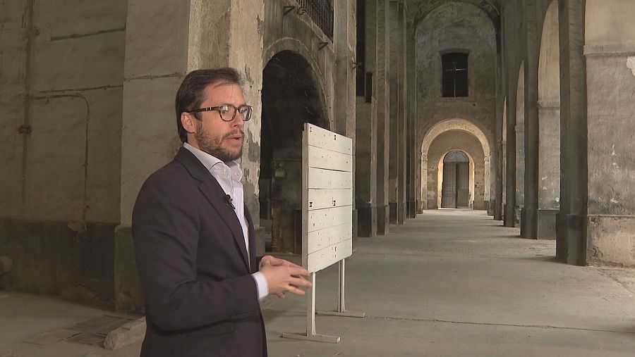 Director de Cultura explica algo en la Real Fábrica de Artillería de Sevilla, junto a un panel informativo en un pasillo con arcos.