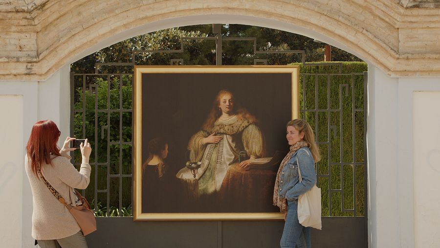 Gran cuadro de Rembrandt expuesto tras verja metálica. Dos mujeres lo observan, una fotografiándolo con su móvil.