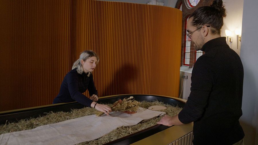 Mujer coloca elementos vegetales sobre una tela en una cápsula dorada, observada por un hombre.  Ambiente cálido y tenue.