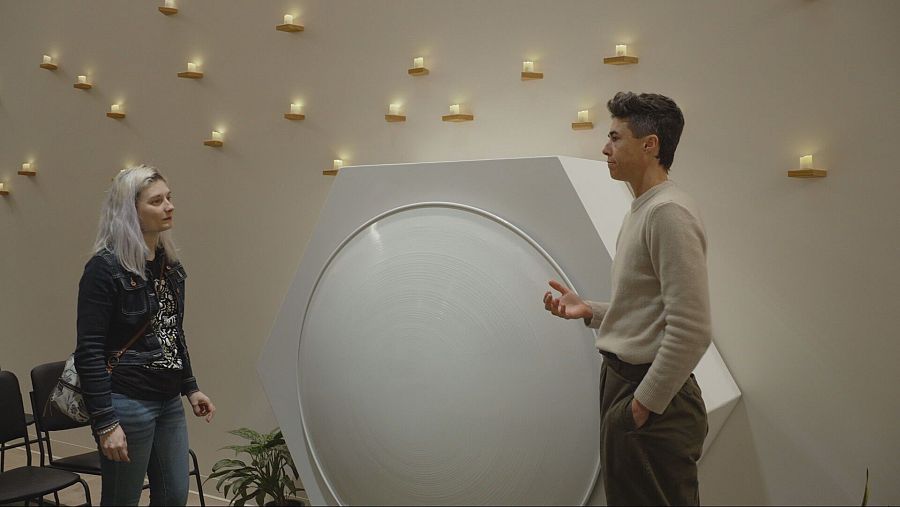 En una sala con velas encendidas, dos mujeres discuten junto a una estructura hexagonal blanca para el compostaje de restos humanos.