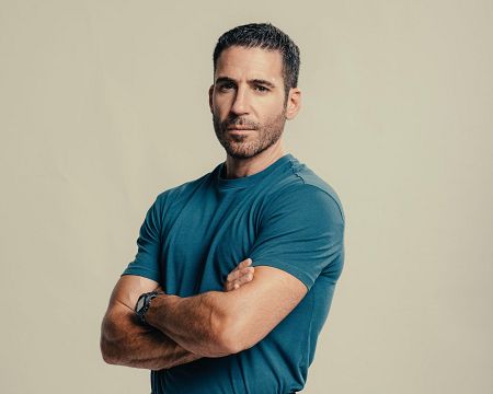 Retrato de un hombre con barba y pelo corto, camiseta azul verdosa y brazos cruzados, proyectando confianza. Fondo beige claro.