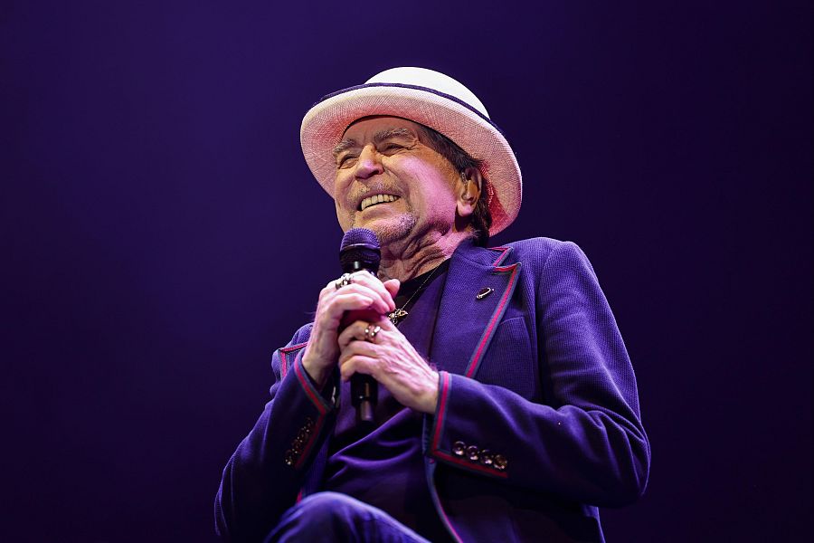Imagen de Joaquín Sabina en concierto: sentado, con micrófono, sombrero claro y chaqueta azul oscura con ribetes granates. Expresión serena.