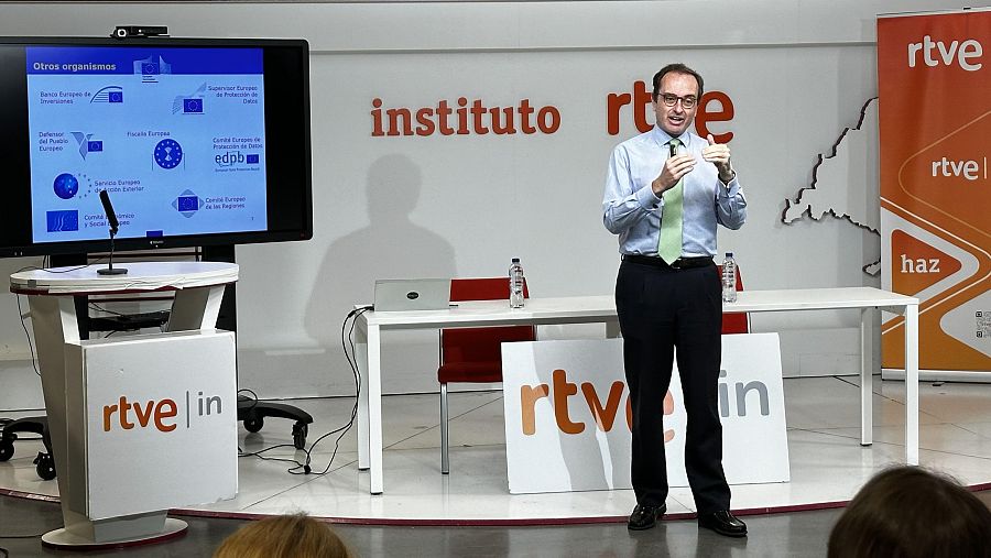 Presentación de Lucas González Ojeda en evento RTVE Instituto.  Gesticula mientras habla ante pantalla con logotipos de organismos europeos.  Asistentes visibles en primer plano.