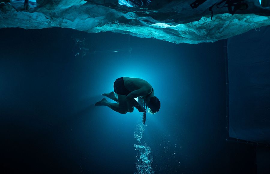 Hombre sumergido en aguas oscuras, encogido y exhalando aire; sobre él, una capa de hielo. Iluminación contrastada en figura.