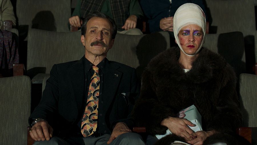 Dos personas en butacas de cine; un hombre en traje oscuro y una persona con abrigo de piel y la cabeza vendada, sujetando un paquete.