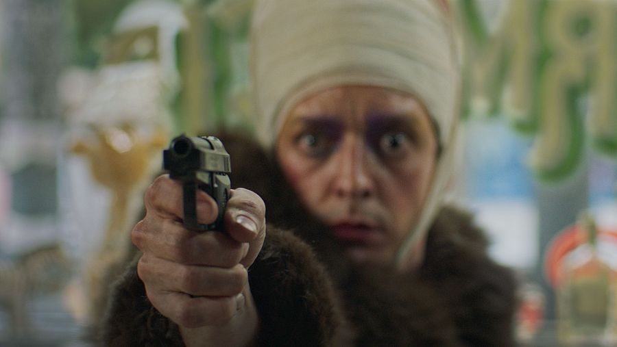 Persona vendada apuntando con una pistola a cámara; expresión tensa; abrigo marrón; fondo interior desenfocado.