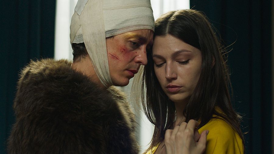 Escena íntima entre un hombre herido, con vendaje en la cabeza y abrigo marrón, y una mujer de pelo oscuro y ropa amarilla.  Momento de vulnerabilidad y cercanía.