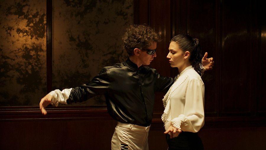 Pareja bailando en un interior elegante, con iluminación tenue. Él lleva camisa negra y pantalones claros; ella, blusa blanca y falda/pantalón oscuro.  Escena de la película 'El Jockey'.