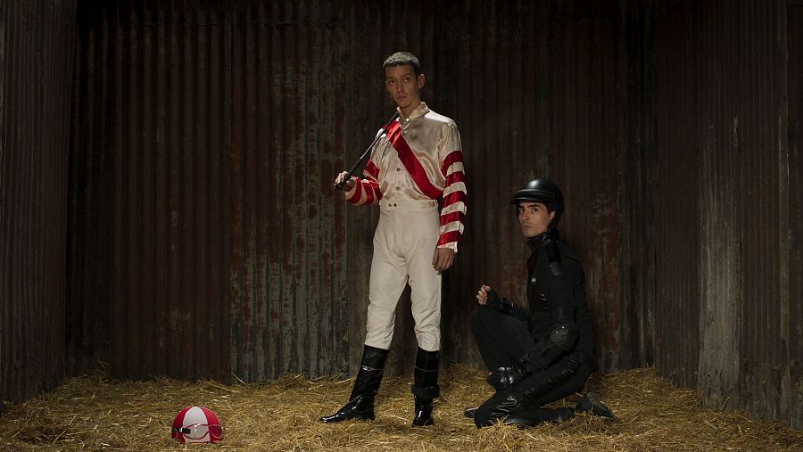 Jockey con chaquetilla blanca y roja y asistente de negro en un establo.  Fusta y casco en el suelo.