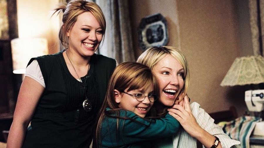 Tres mujeres sonríen en una escena interior; una niña abraza a una de ellas.  Ambiente hogareño, con una lámpara y una máquina de coser al fondo.