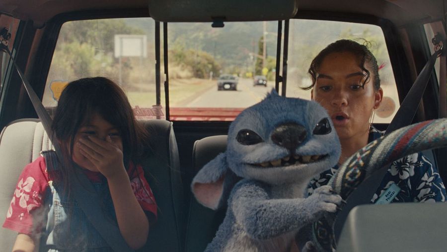 Escena en un coche: una niña se esconde, una joven parece preocupada y una criatura azul parece estar causando problemas al volante.