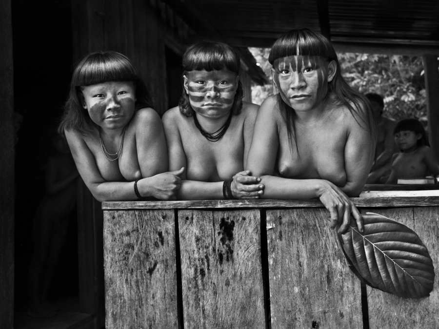 Tres mujeres indígenas amazónicas, con pintura facial y collares, posan en una estructura de madera.  Imagen en blanco y negro con una atmósfera serena.