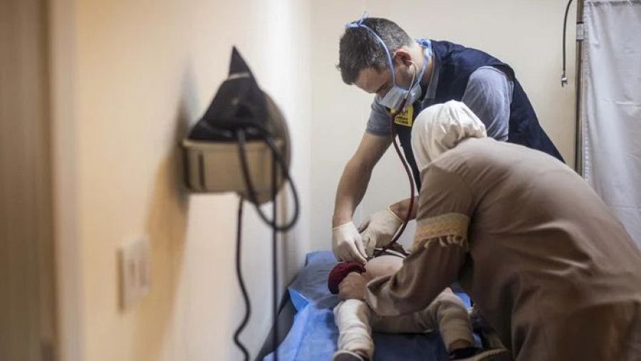 Médico examina a un niño con estetoscopio; una mujer lo acompaña.  Consulta médica sencilla con tensiómetro visible.