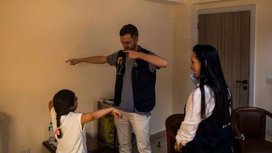 Hombre con estetoscopio explica a una niña; mujer observa sonriendo.  Escena en habitación con muebles sencillos, posible contexto educativo o de salud.