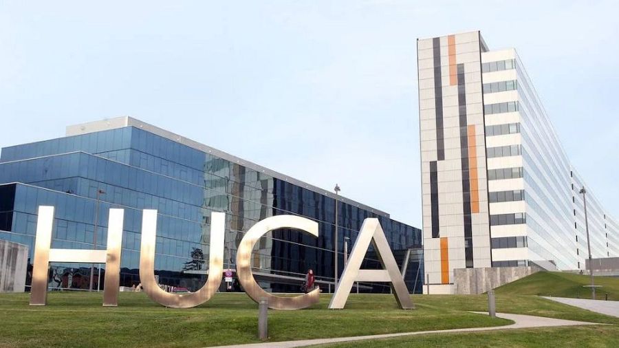 Imagen del HUCA: letras metálicas en césped, edificio principal con fachada de cristal azul y torre blanca con franjas verticales.